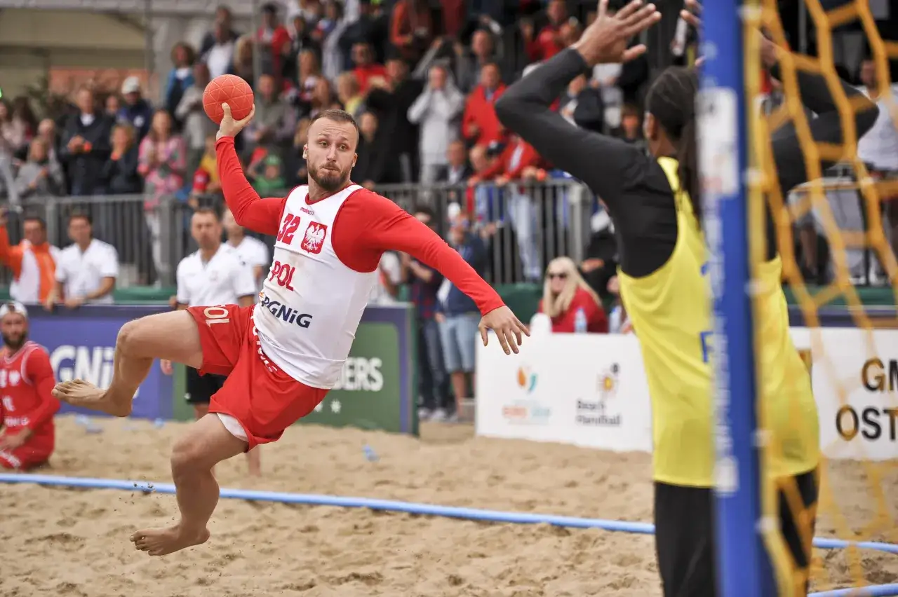 Filip Jankowski w skoku rzuca piłką do bramki podczas meczu piłki ręcznej plażowej.