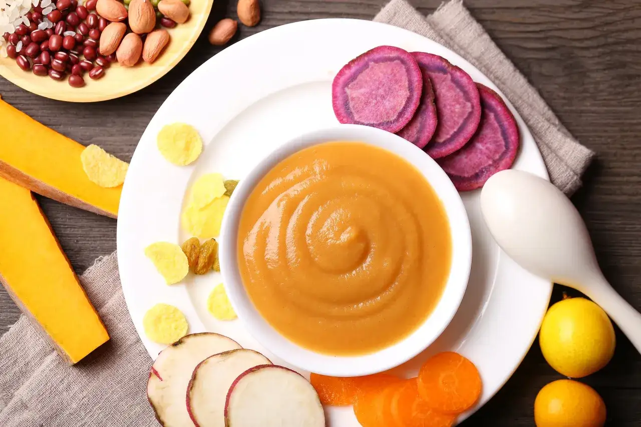 Pyszna, kremowa **kolacja dla niemowlaka** z dyni, batatów i marchewki. Obok miseczki z puree leżą plasterki fioletowego batata, kawałki dyni, marchewki, kukurydziane płatki i rodzynki.