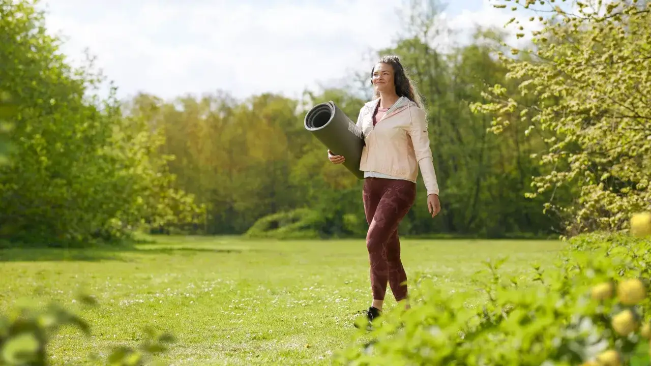 Vrouw met sportmat en koptelefoon loopt in park. Ze lijkt last te hebben van voorjaarsmoeheid symptomen, maar zoekt ontspanning in de natuur.