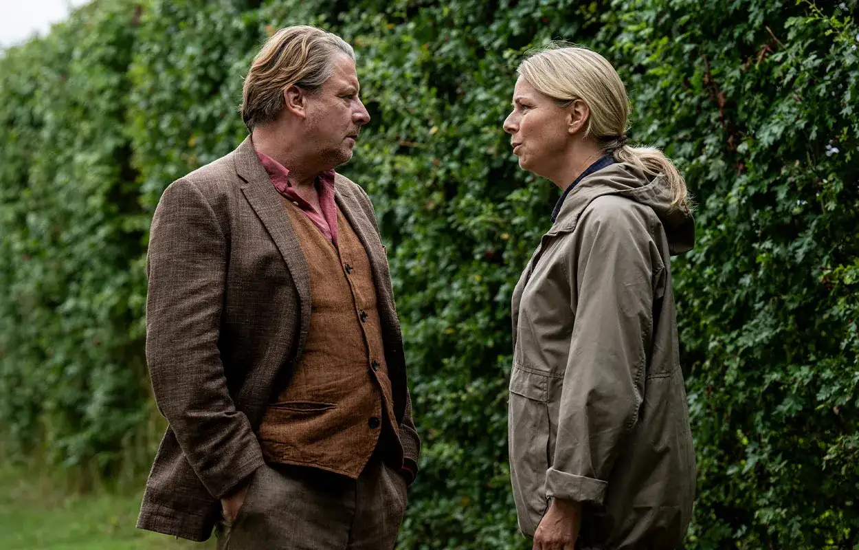 Två personer står och pratar vid en grön häck. Mannen har kavaj och väst, kvinnan en grön jacka. En scen från en ny dansk serie på TV4.