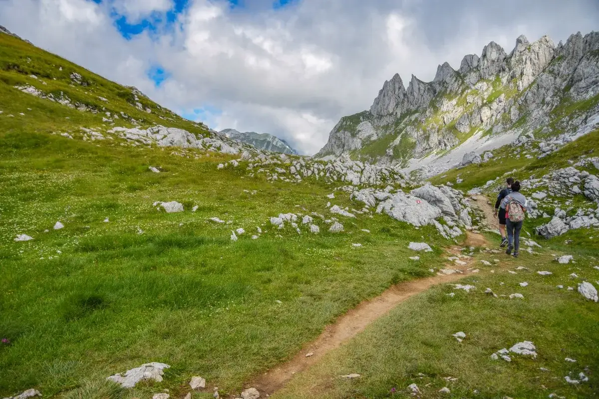 Wanderer erkunden einen Pfad im Durmitor Nationalpark. Grüne Hänge, Felsen und dramatische Berggipfel unter blauem Himmel.