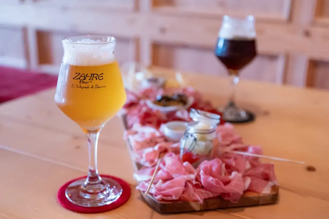 Un bicchiere di birra Zahre dorata, con schiuma abbondante, accanto a un tagliere di salumi e un altro bicchiere di birra scura.