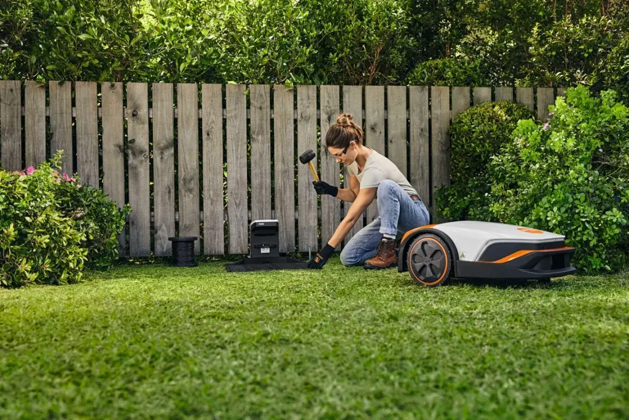 Kobieta instaluje stację dokującą, przygotowując teren do pracy robota koszącego. Pokazuje, jak działa robot koszący.