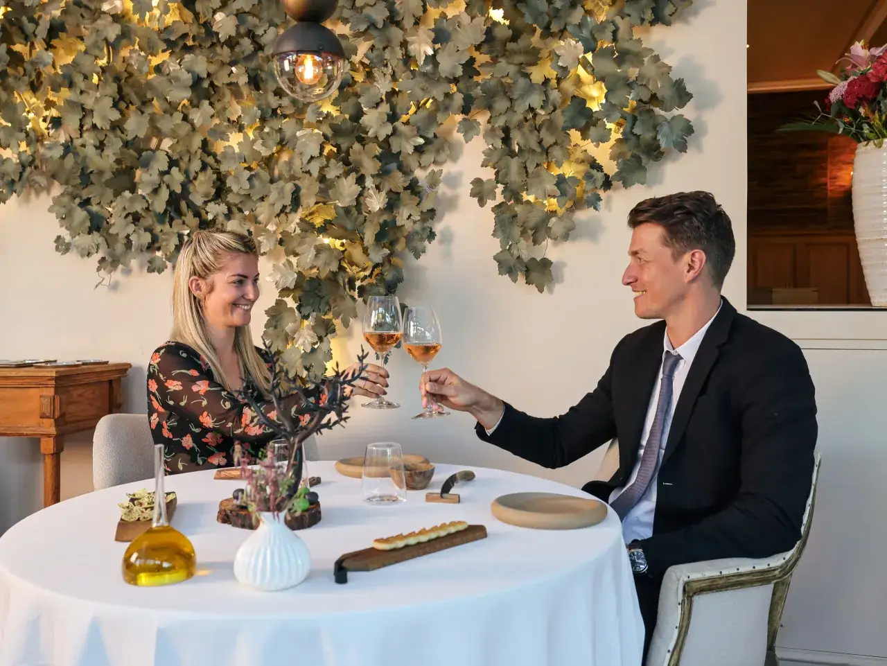 Un couple trinque avec des verres de ros&eacute; dans un restaurant gastronomique, d&eacute;cor&eacute; de feuilles stylis&eacute;es.