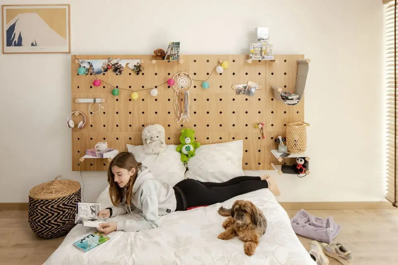 Une jeune fille lit sur son lit, un chien &agrave; ses c&ocirc;t&eacute;s. La t&ecirc;te de lit en bois perfor&eacute; est d&eacute;cor&eacute;e de photos, de guirlandes lumineuses et d'objets. Id&eacute;e pour comment fixer t&ecirc;te de lit.