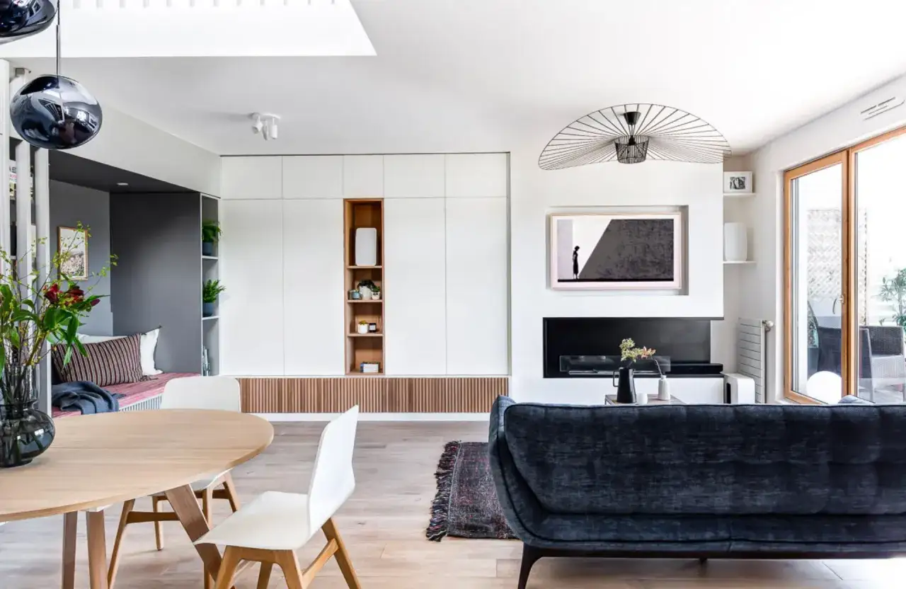 Un salon moderne et lumineux avec une table en bois, des chaises blanches, un canap&eacute; bleu nuit et une grande armoire murale. La d&eacute;coration sugg&egrave;re un grand taille salon.