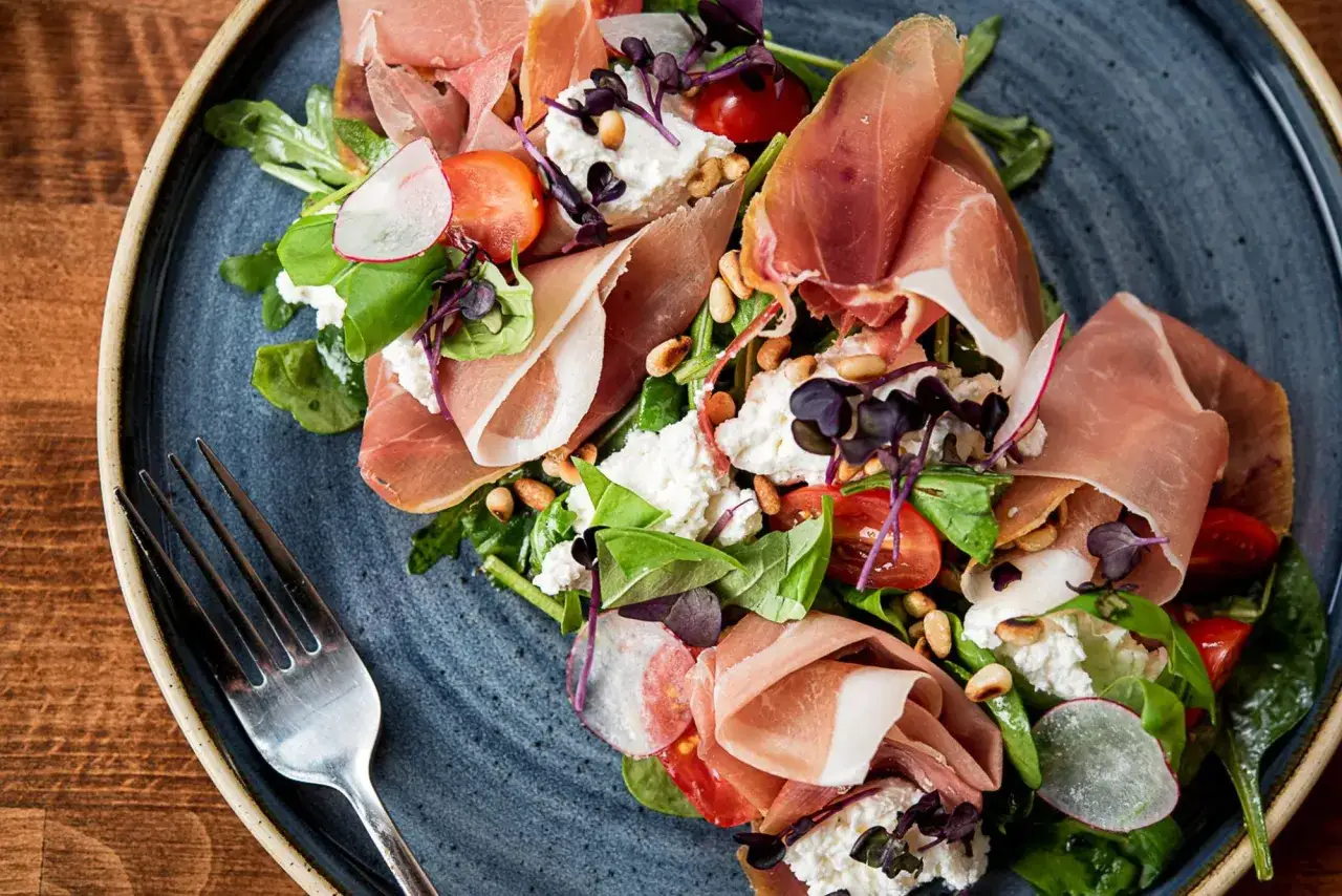 Wykwintna kolacja po włosku: prosciutto, ser ricotta, rukola, pomidorki koktajlowe i orzeszki piniowe na talerzu.