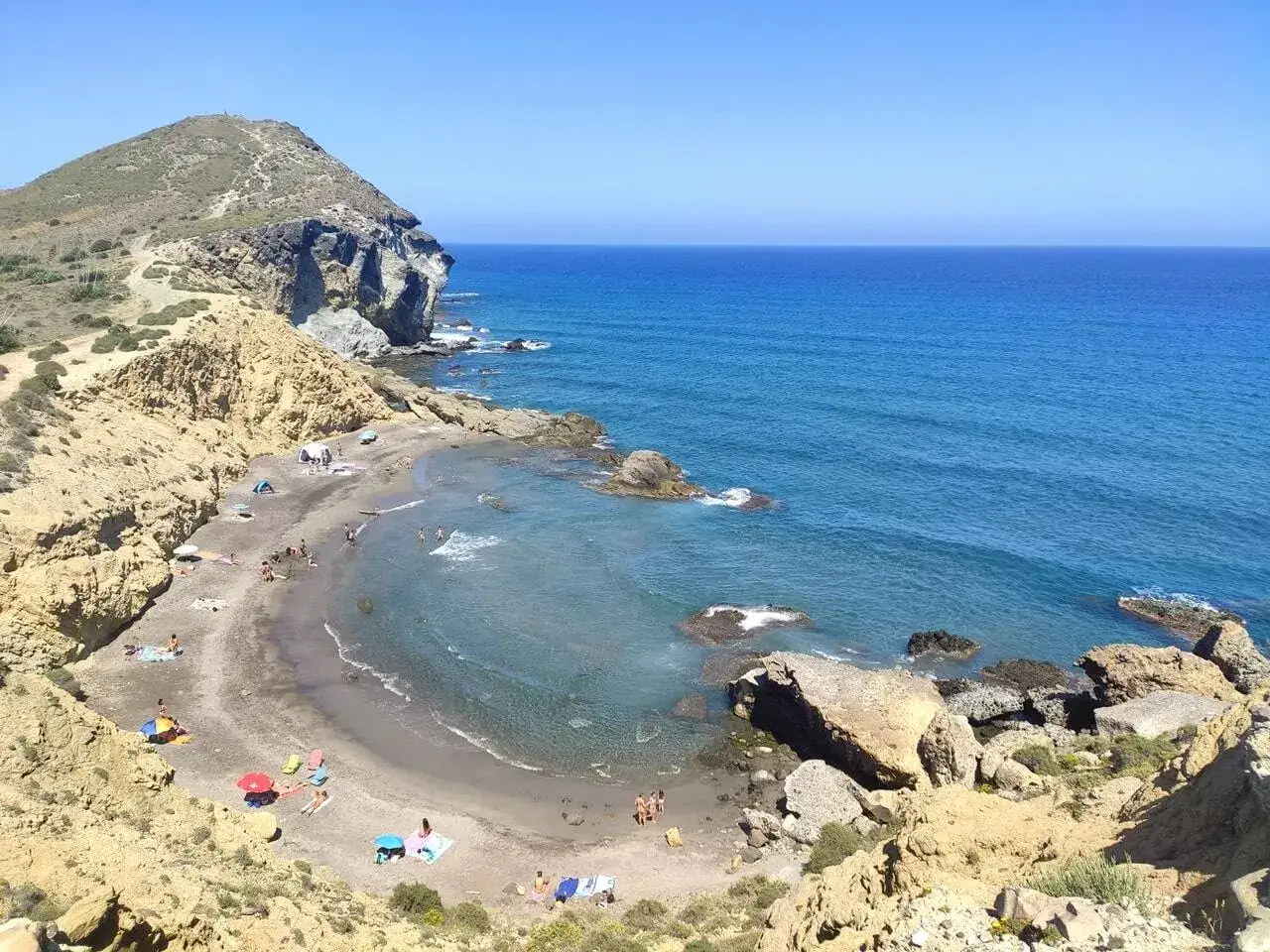 Vista panorámica de la cala cortada en Almería, con gente disfrutando de la playa y el mar azul bajo un cielo despejado.