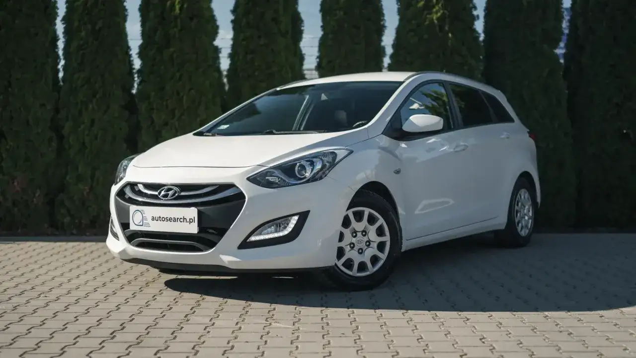 Biały Hyundai i30 1.4 benzyna, kombi, zaparkowany na kostce brukowej przed żywopłotem.