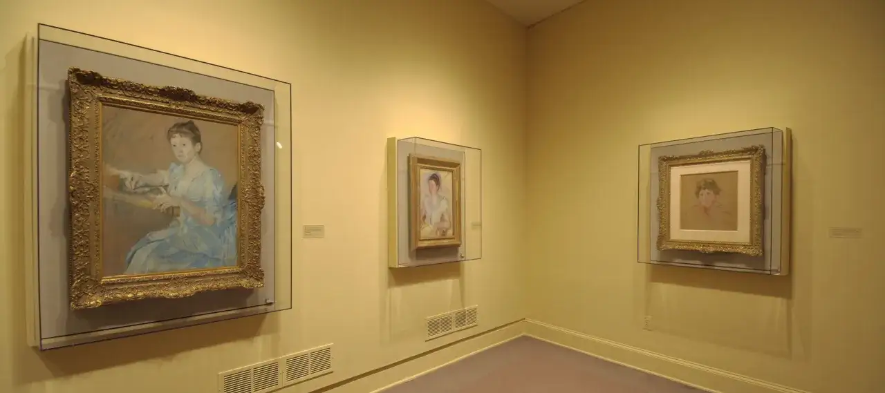 Three framed portraits, likely Mary Cassatt paintings, hang on a pale yellow wall in a gallery.