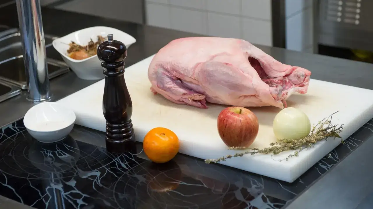 Eine rohe Gans liegt auf einem Schneidebrett, bereit zum Auftauen. Daneben liegen ein Apfel, eine Zwiebel, eine Orange und ein Pfeffermühlen.