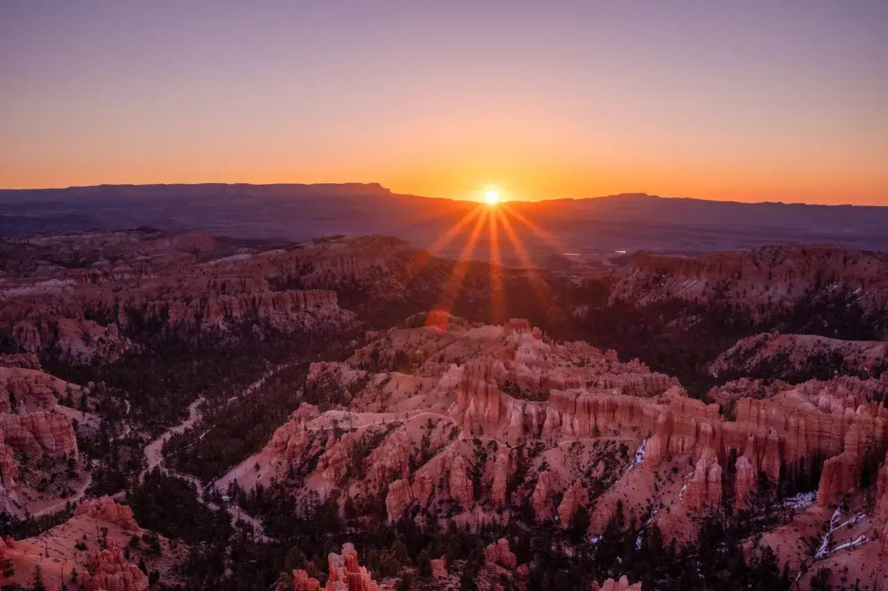 Zachód słońca nad Bryce National Park, malujący niebo od fioletu po pomarańcz. W dole rozciągają się skalne iglice i wąwozy.