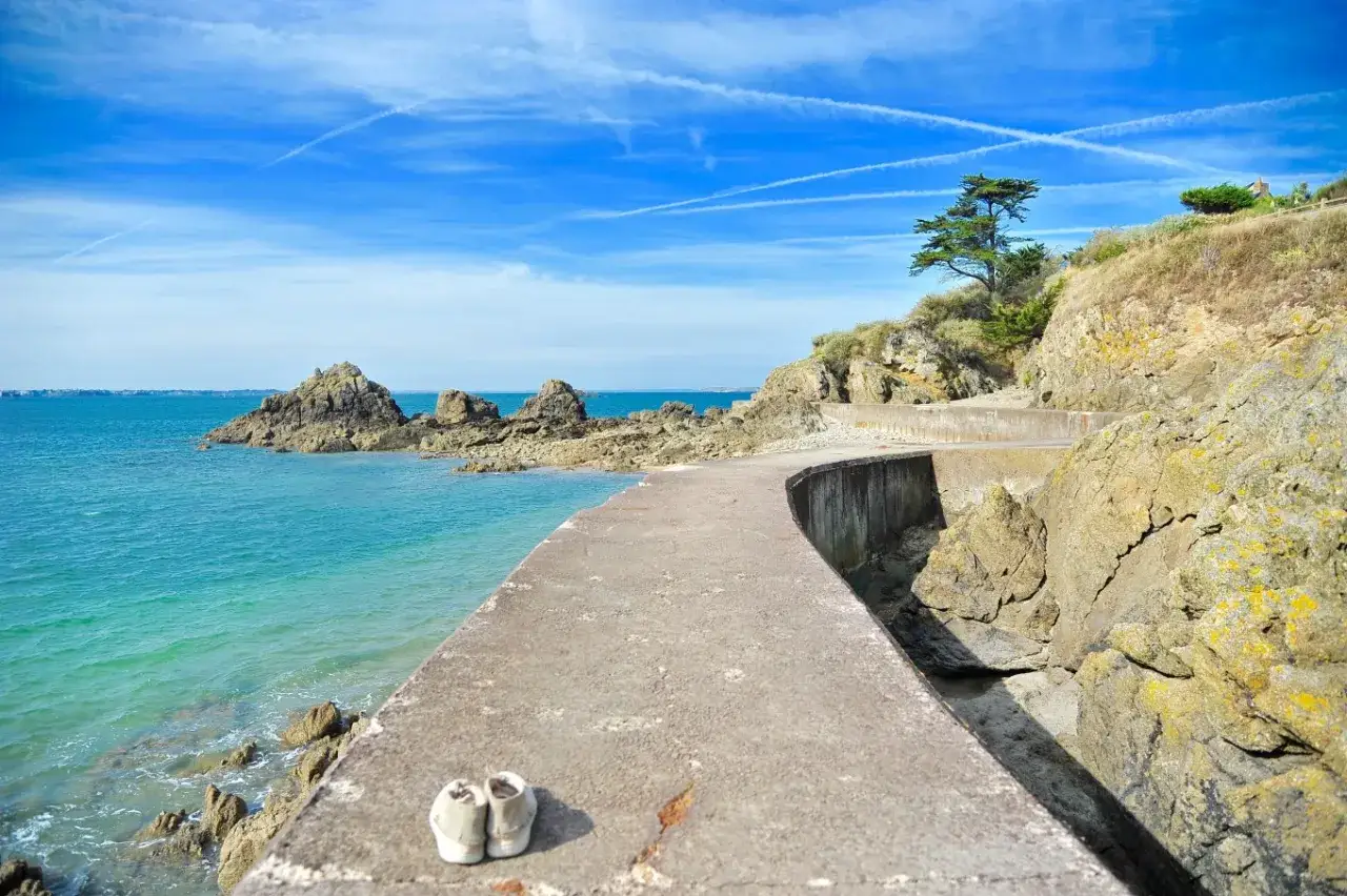 Une paire de chaussures abandonnée sur le chemin des douaniers à Saint-Malo, face à la mer turquoise et aux rochers.
