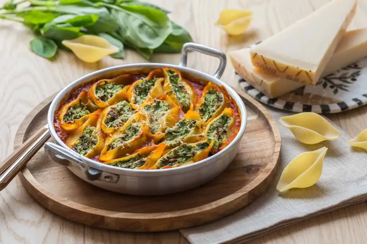 Un plat de conchiglioni farcis, garni de fromage r&acirc;p&eacute;, baignant dans une sauce tomate. Des p&acirc;tes et du parmesan sont dispos&eacute;s autour.