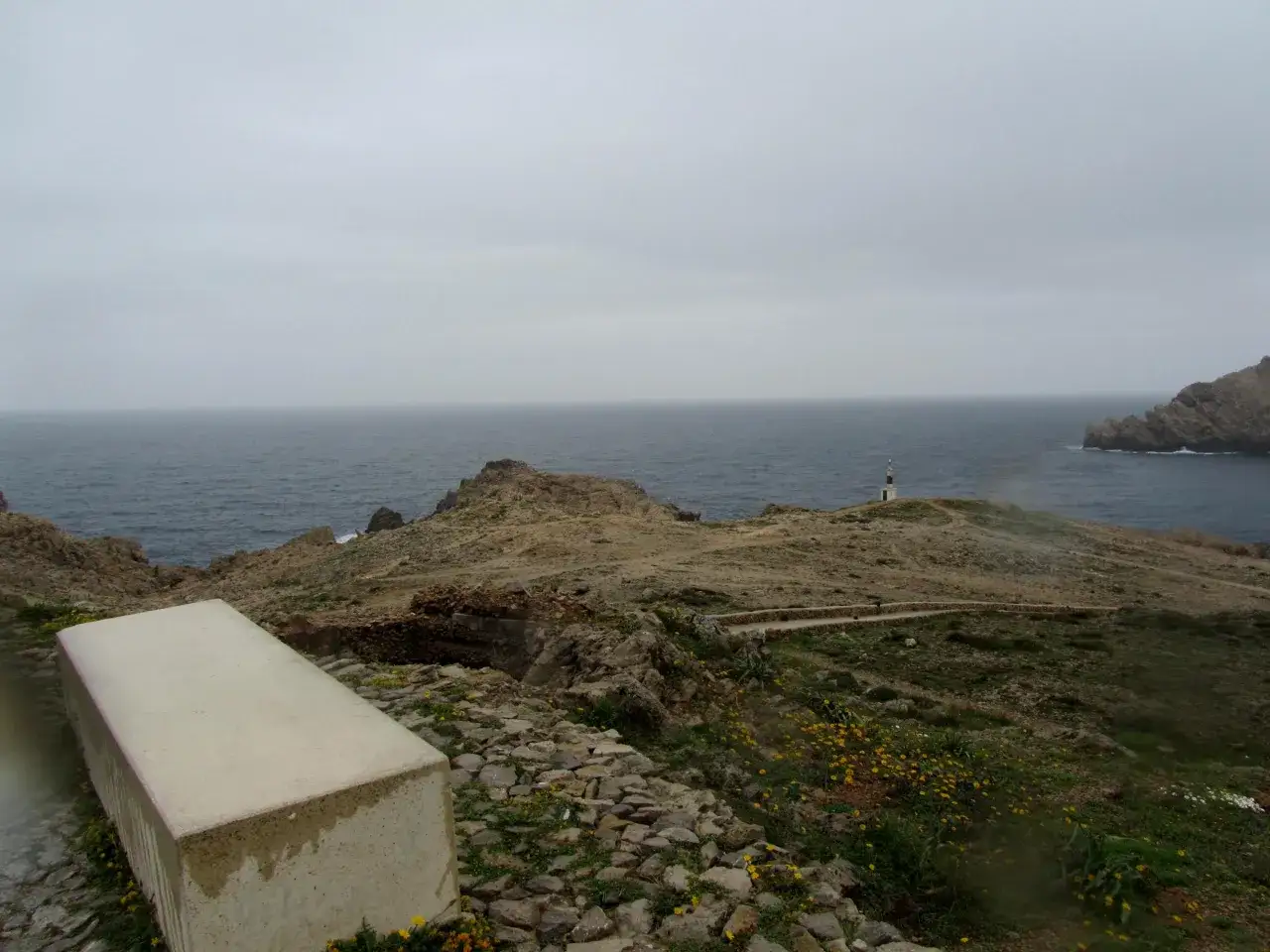 Paisaje costero con un faro al fondo. Un camino serpentea entre rocas y flores amarillas. Ideal para saber qué ver en Fornells.