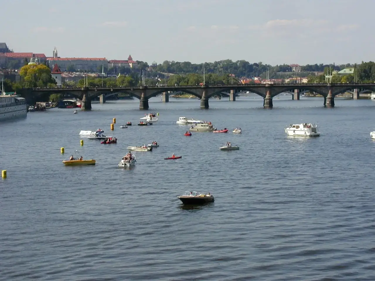 Bateaux et cano&euml;s naviguent sur la Vltava, sous un pont, avec la ville de Prague en arri&egrave;re-plan. Une sc&egrave;ne de 