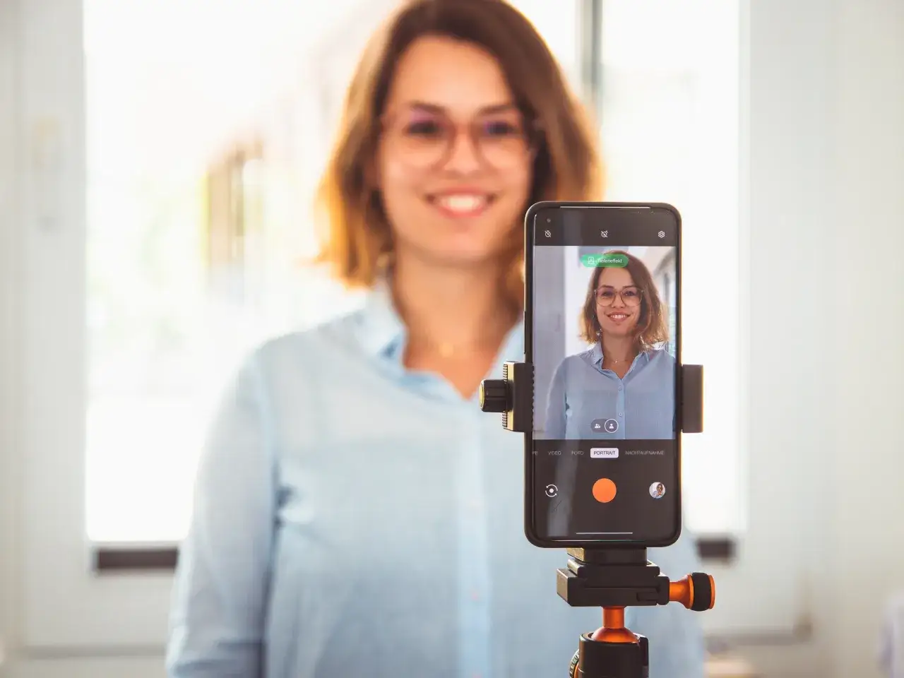 Frau macht ein bewerbungsfoto selber machen mit dem Handy auf einem Stativ. Sie l&auml;chelt freundlich in die Kamera.