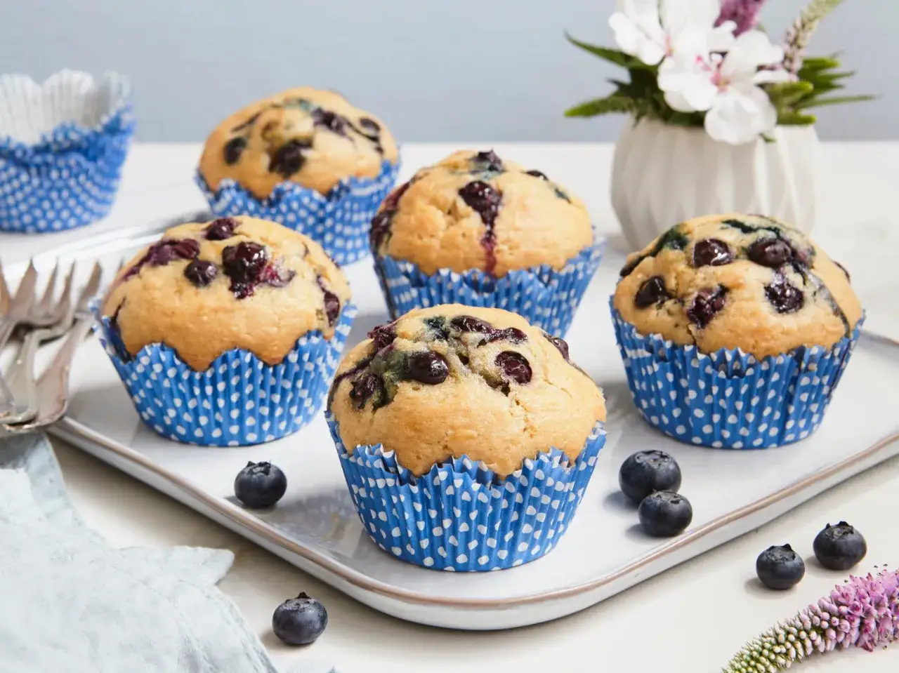 Fünf saftige, vegane Blaubeermuffins in blauen Papierförmchen auf einem Teller, garniert mit frischen Blaubeeren und einer Blume.