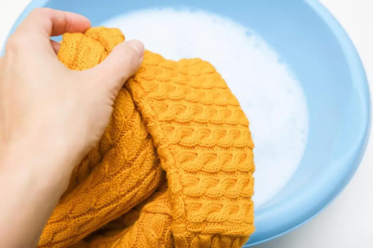 Ręka trzyma żółty sweter nad niebieską miską z wodą i pianą. Delikatne pranie pomoże uniknąć mechacenia.