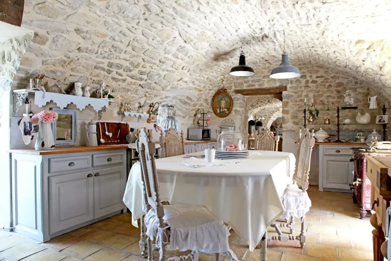 Cuisine au style shabby chic avec murs en pierre, table dress&eacute;e et vaisselle ancienne.