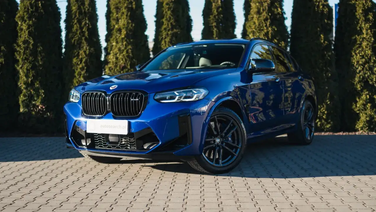Błękitny suv BMW X4 M Competition lśni w słońcu, zaparkowany na kostce brukowej przed żywopłotem.