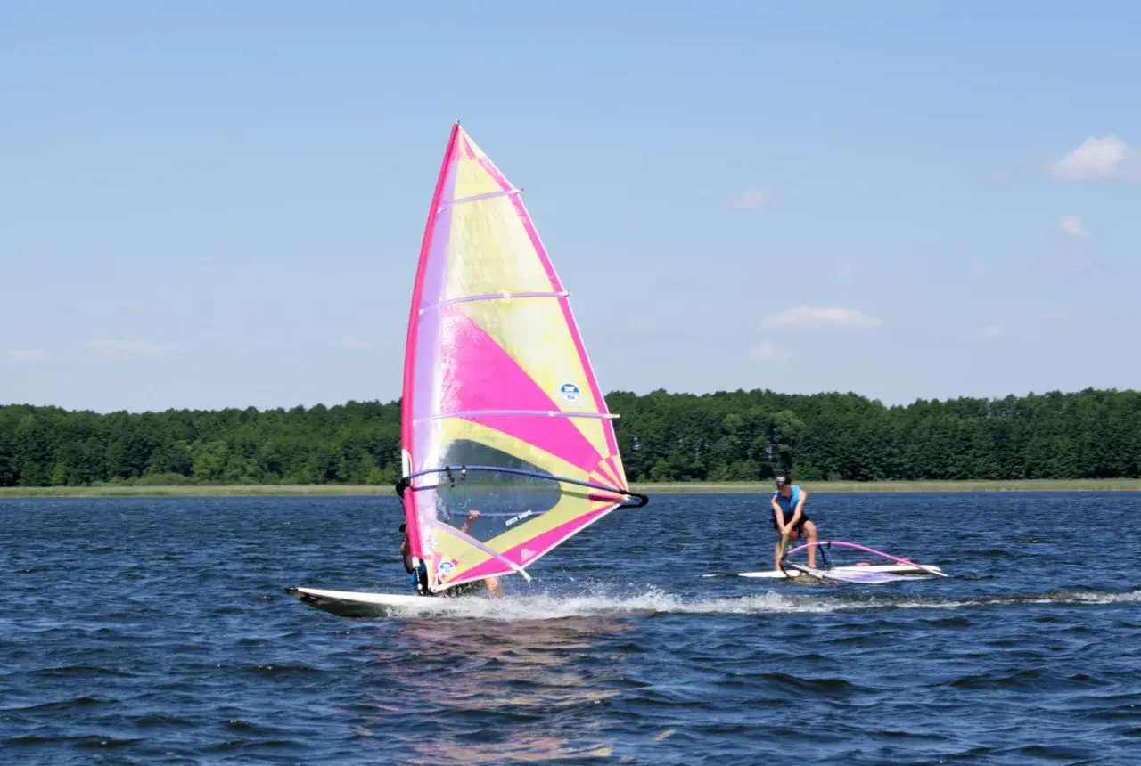 Dwóch surferów na Mazurach, jeden z kolorowym żaglem, cieszy się wiatrem i wodą. Idealne miejsce na windsurfing Mazury.