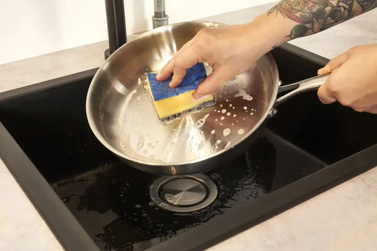 Hand reinigt eine Edelstahlpfanne mit einem Schwamm in der Sp&uuml;le. So wird die beschichtete pfanne reinigen.