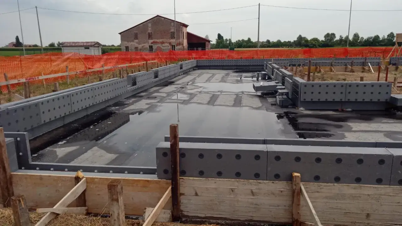 Costruzione di una platea di fondazione con blocchi prefabbricati grigi. L'area è parzialmente allagata.