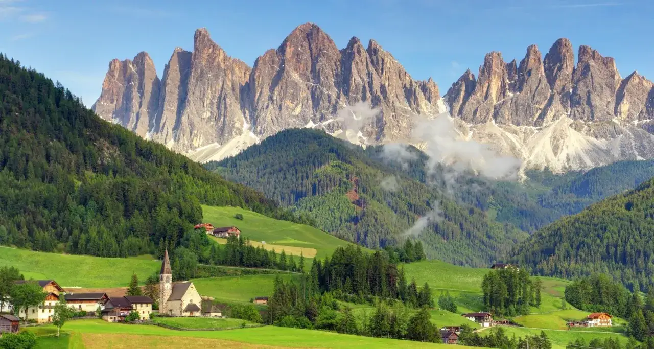 Un pueblo alpino con una iglesia y casas se asienta en un valle verde, con bosques y picos imponentes de las Dolomitas al fondo. Ideal para trekking.