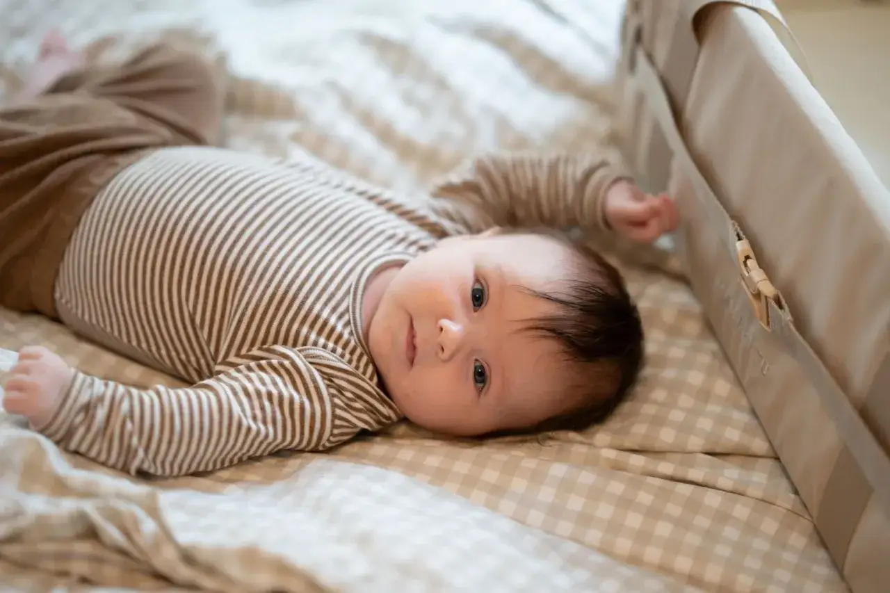 En 1 m&aring;nads bebis sover inte, utan ligger vaken p&aring; en randig body och bruna byxor. Bebisen tittar mot kameran.