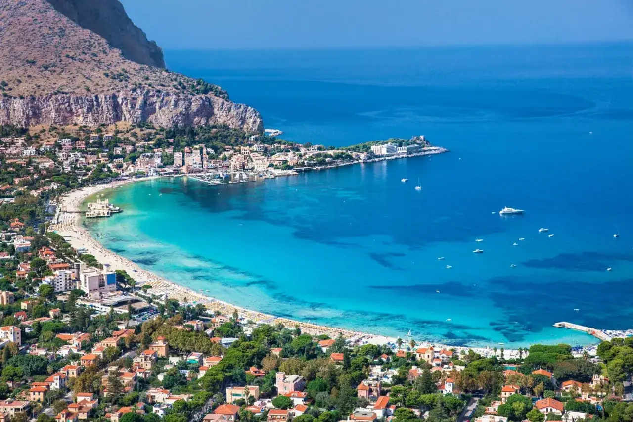 Vista aerea di una baia con acqua turchese, spiaggia affollata e case colorate. Il mare &egrave; di un blu intenso, quasi viola come il mare, con barche a vela e yacht.
