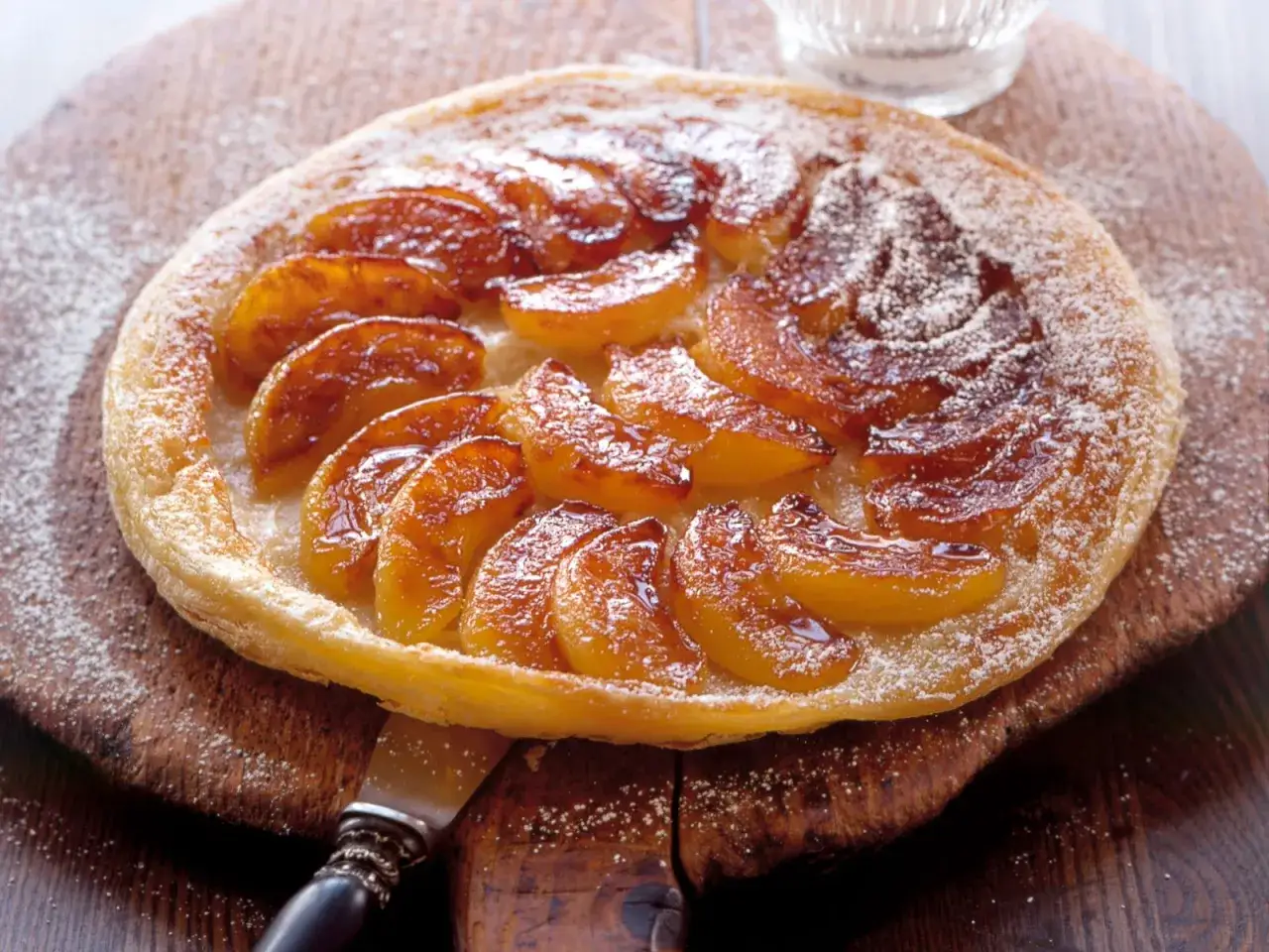 Eine karamellisierte Tarte Tatin mit saftigen Apfelspalten, best&auml;ubt mit Puderzucker, auf einem rustikalen Holzbrett.