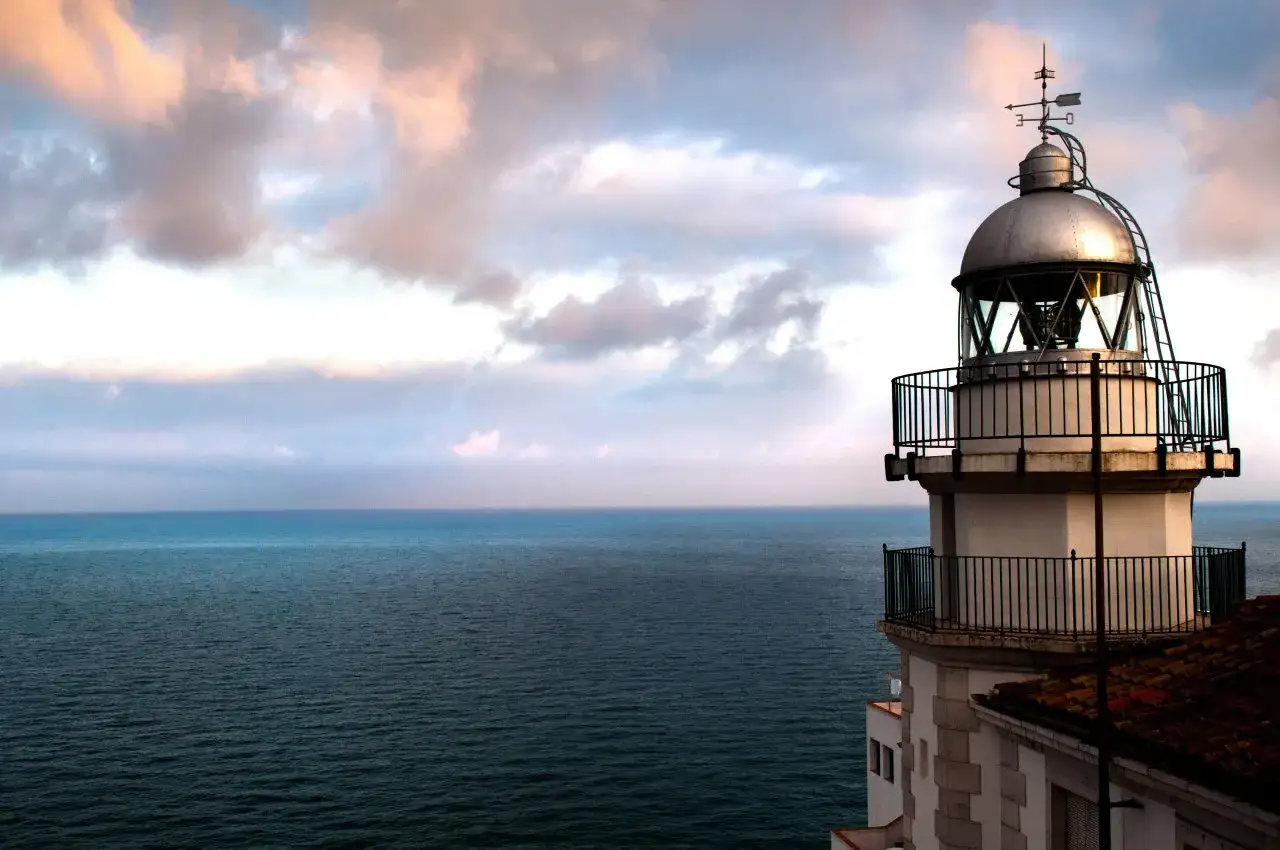 El faro de Pe&ntilde;&iacute;scola se alza sobre el mar azul bajo un cielo de nubes rosadas y grises.