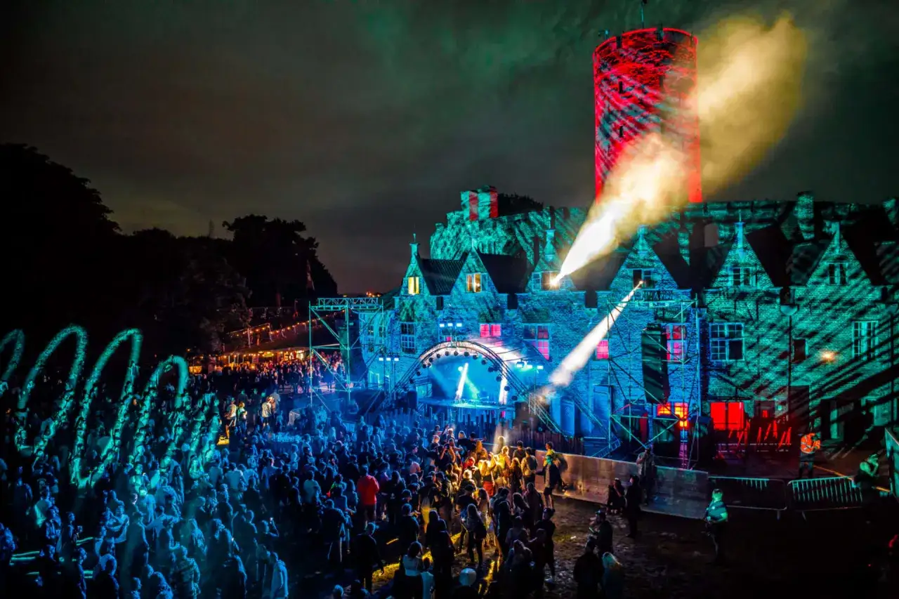 Nocny festiwal techno w Polsce. Tłum bawi się przy scenie oświetlonej kolorowymi światłami, a zamek w tle rozświetlają projekcje.