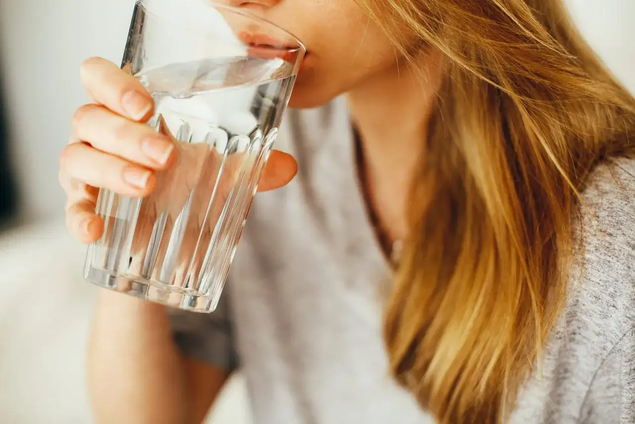 Une femme aux cheveux blonds boit un verre d'eau. Une question importante pour les jeunes mamans : quelle eau boire allaitement ?