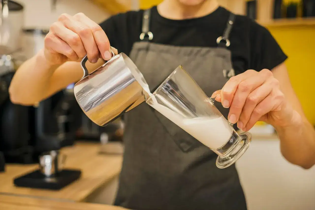 Barista wylewa spienione mleko z dzbanka do szklanki, pokazując, jak zrobić latte.