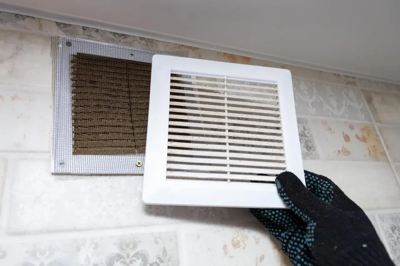 Guante quita rejilla de ventilación sucia, mostrando filtro con moho. Busca remedios caseros para quitar el moho de las paredes.