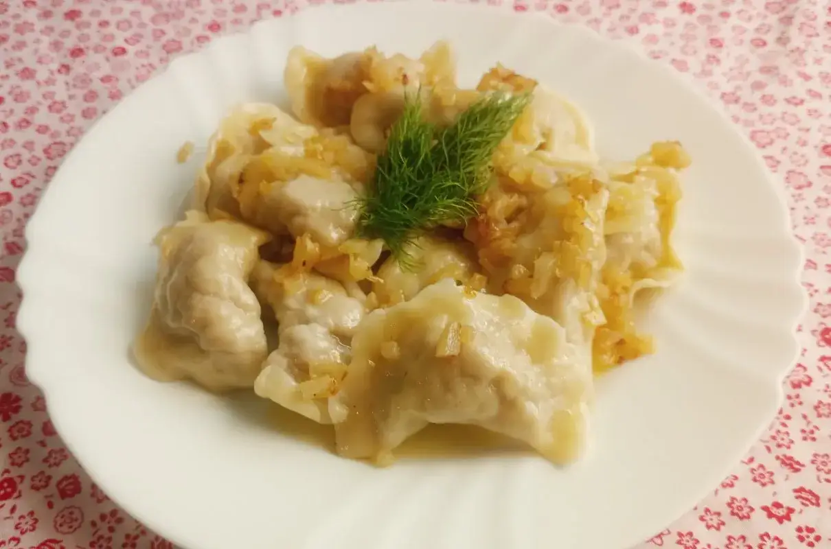 Pyszne pierogi z mięsem, polane skwarkami z cebulki i ozdobione gałązką koperku, prezentują się apetycznie na białym talerzu.