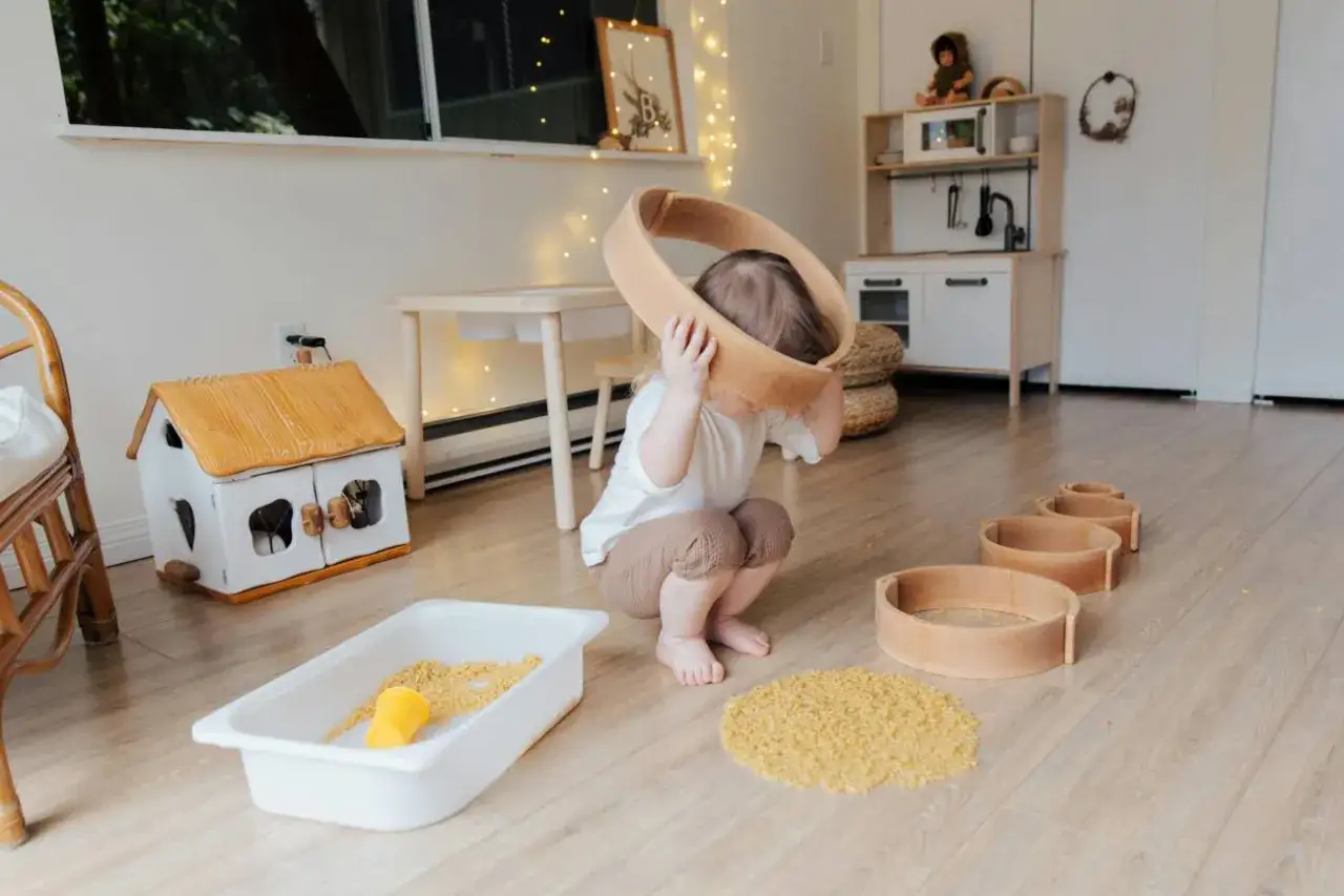 Niña jugando con aros de madera en una **montessori guarderia**. Hay una casita de juguete, una bandeja con arroz y una cocinita de juguete al fondo.