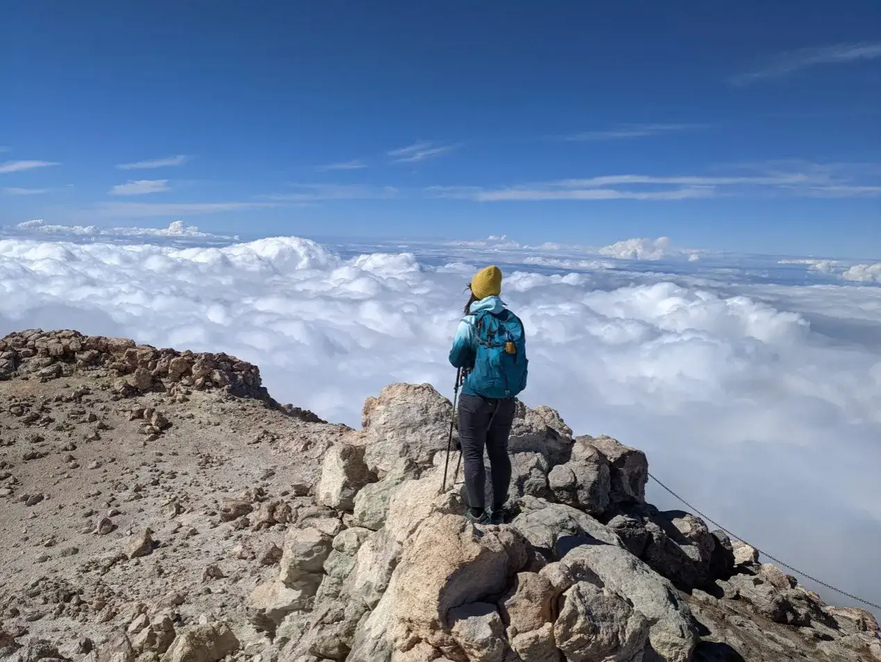Widok z g&oacute;ry na chmury podczas wejścia na Teide. Turystka z plecakiem i kijkami stoi na skalistym szczycie.