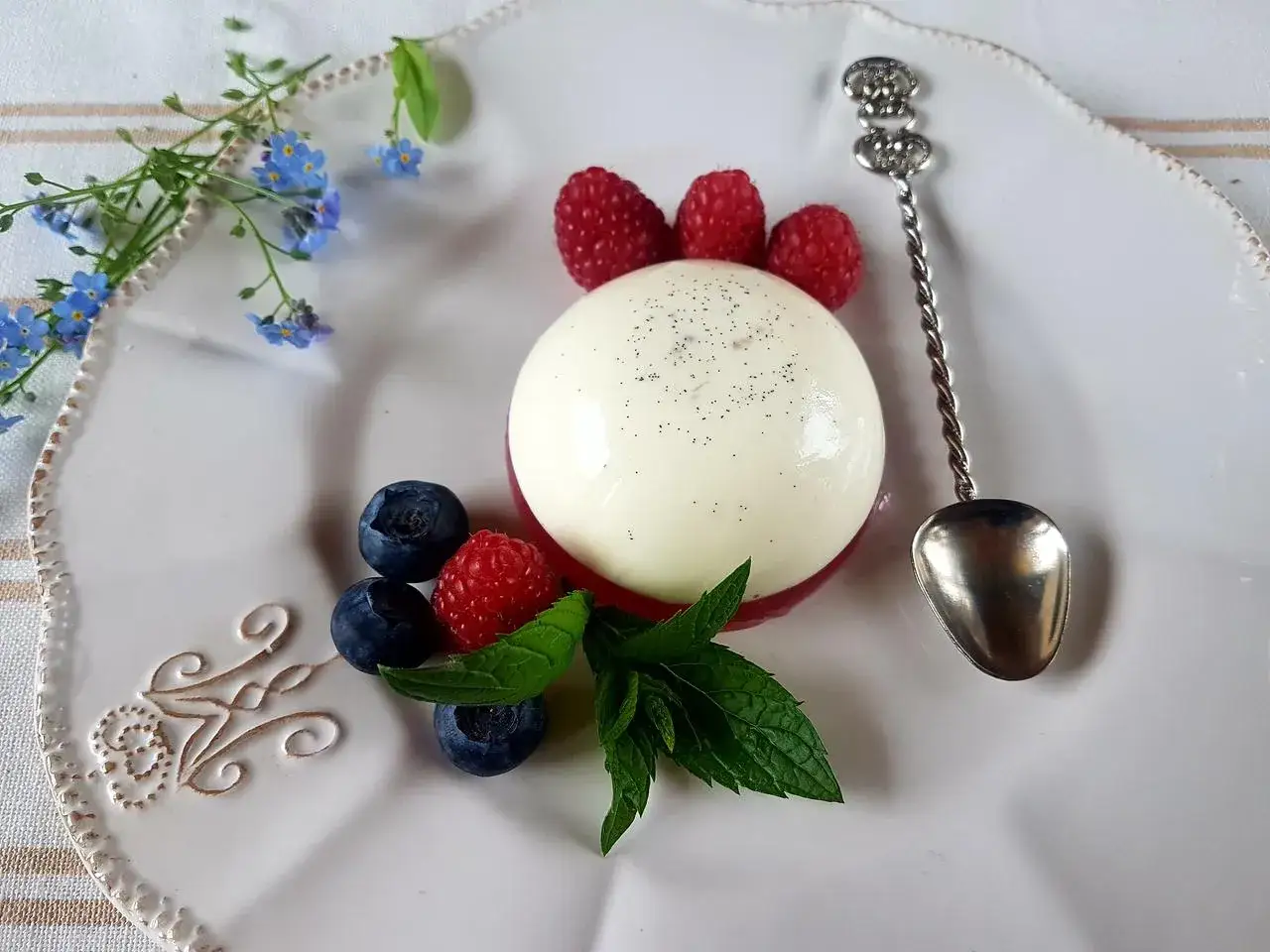 Panna cotta oryginalny przepis: kremowy deser z malinami, borówkami i miętą, ozdobiony kwiatami niezapominajki.