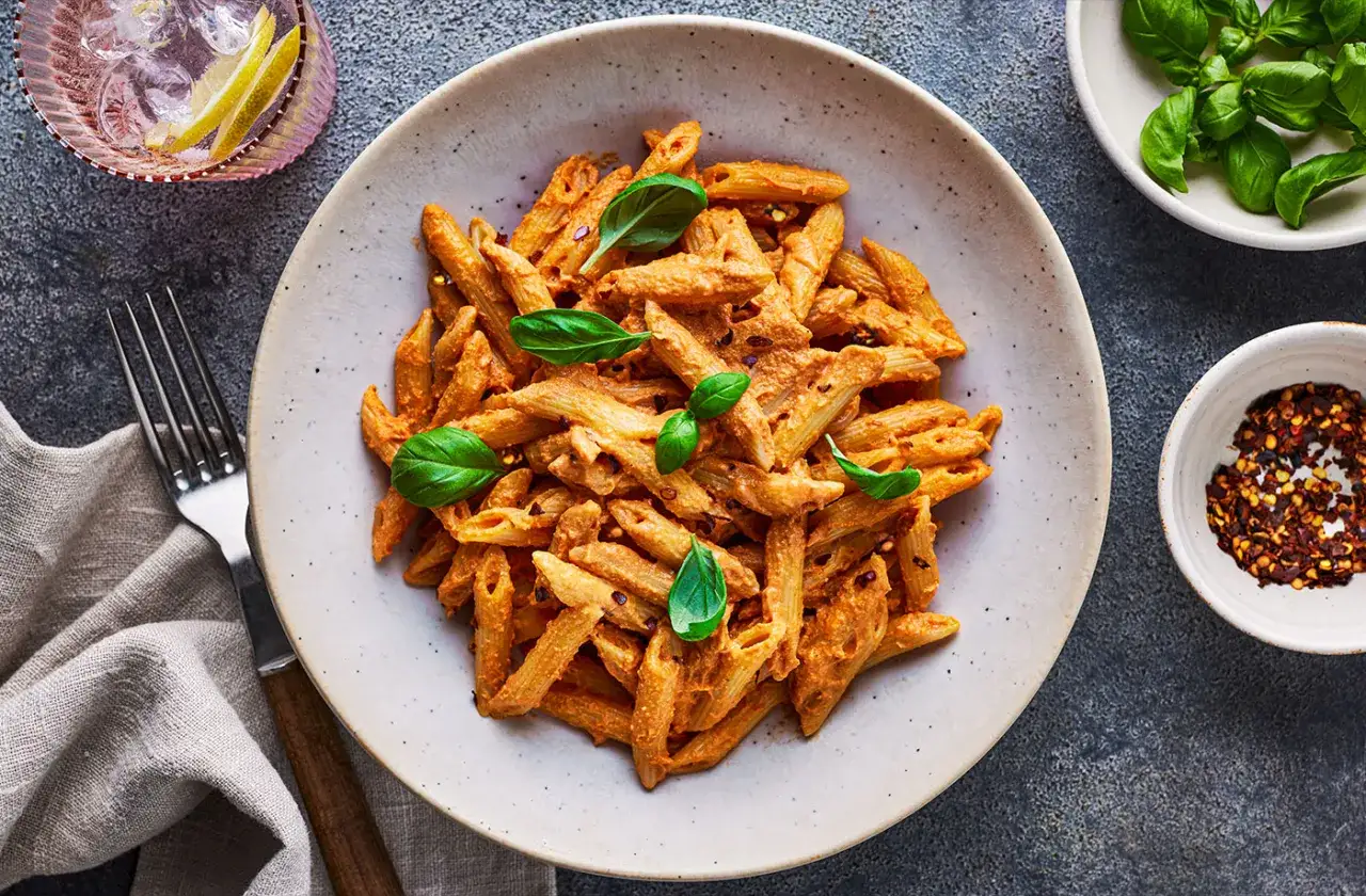 Penne z kremowym sosem pomidorowym i tofu, udekorowane bazylią. Obok miseczka z płatkami chili i szklanka z drinkiem.