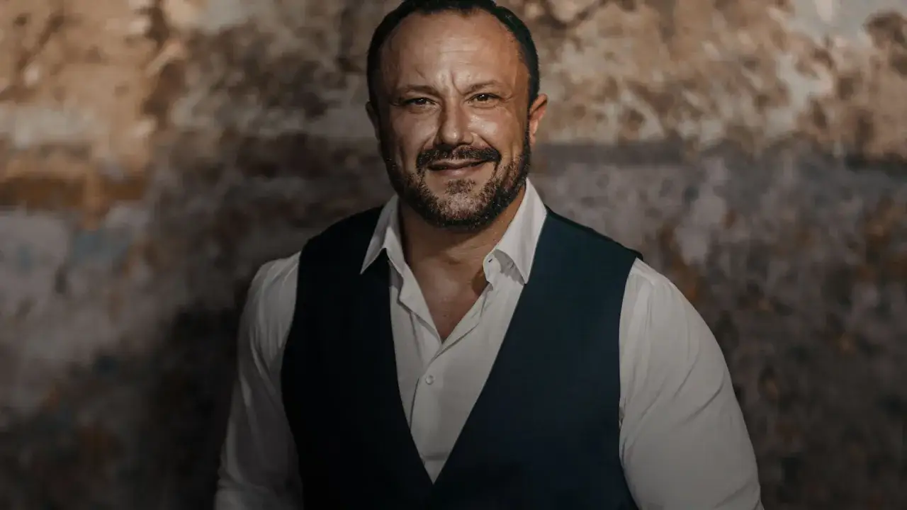 Mirosław Niewiadomski, tenor, w białej koszuli i kamizelce, uśmiecha się na tle rustykalnej ściany.