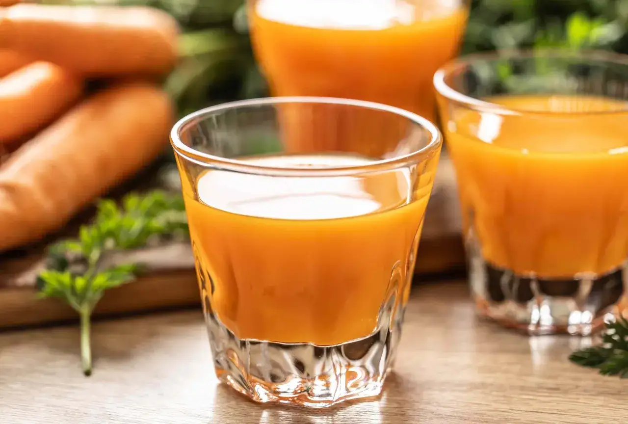 Frisch gepresster Karottensaft in Gläsern, daneben Karotten und Grünzeug. Die positive karottensaft wirkung für die Gesundheit ist bekannt.