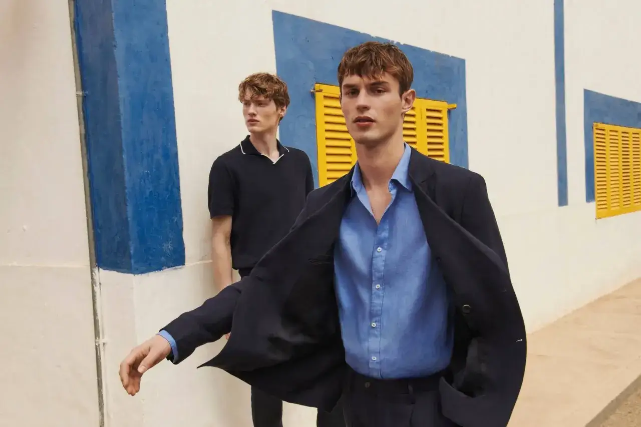 Dos hombres posan con un outfit oficina verano hombre. Uno lleva un traje azul marino y camisa azul claro, el otro un polo oscuro.