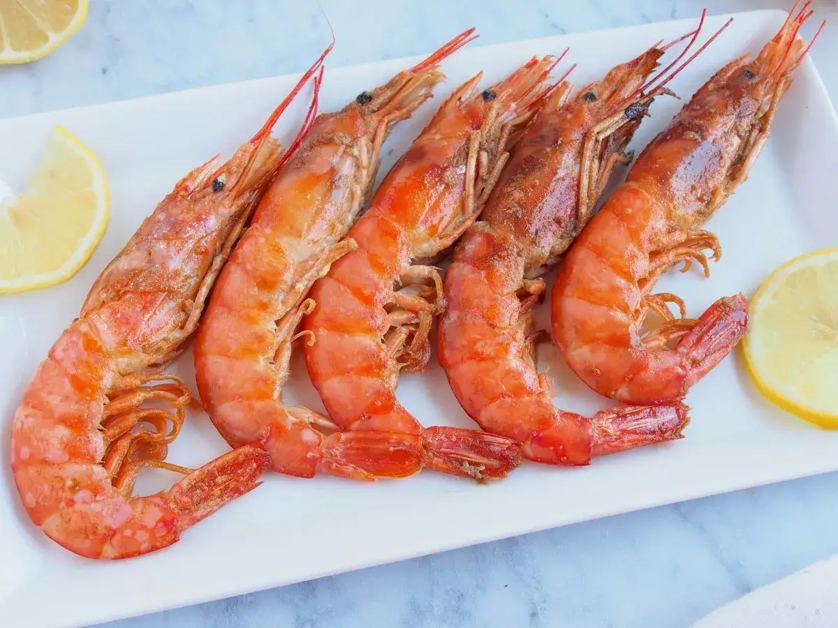 Cinco gambas a la plancha, jugosas y doradas, servidas en plato blanco con rodajas de limón.