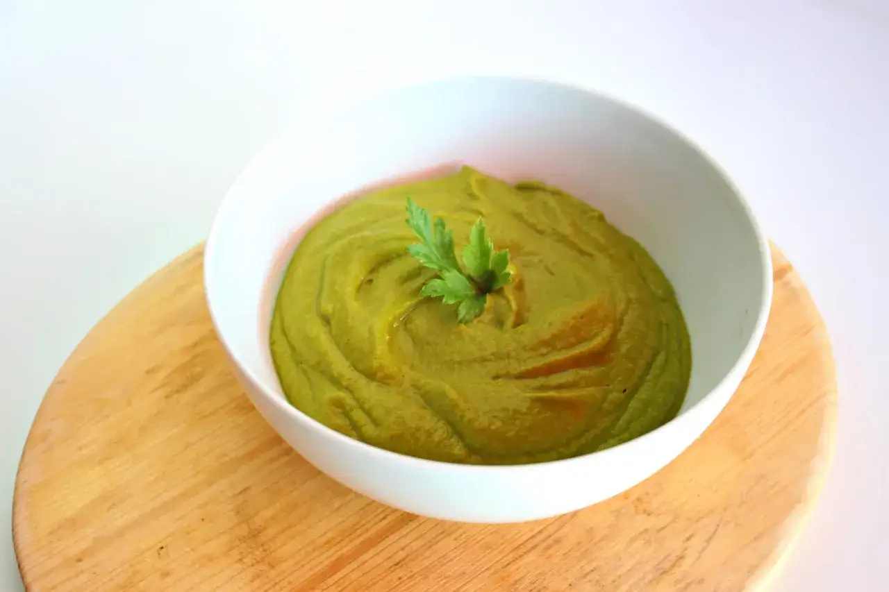 Un bol blanc rempli de pur&eacute;e de brocolis onctueuse, garni d'une feuille de persil frais, pos&eacute; sur une planche en bois.