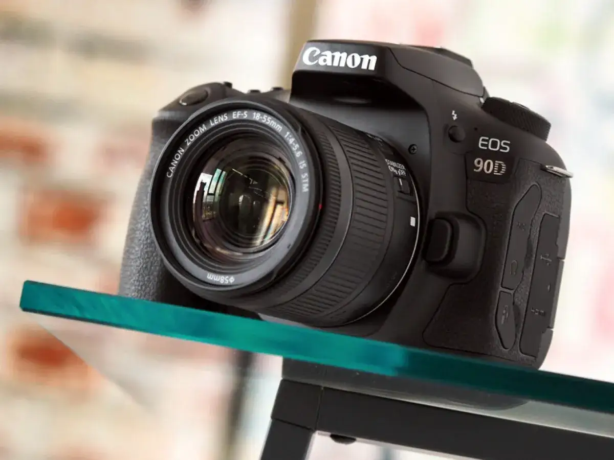 Czarny aparat Canon EOS 90D z obiektywem EF-S 18-55mm stoi na szklanej półce.