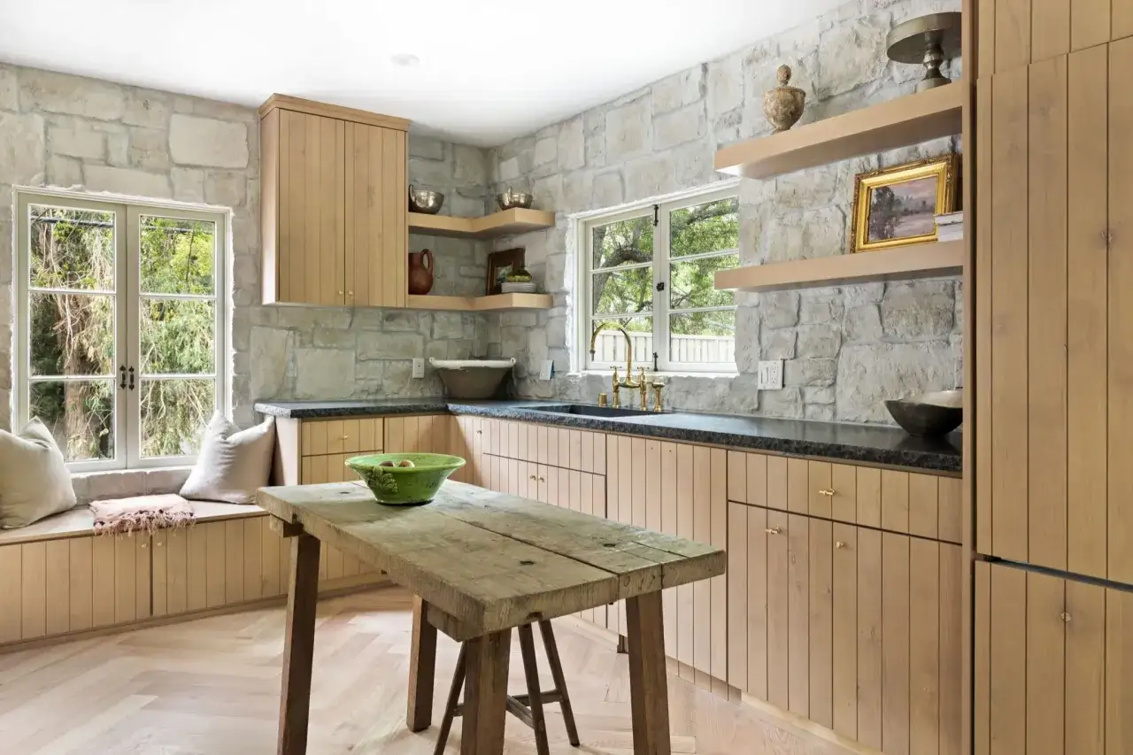 Cocinas con paredes de piedra r&uacute;stica, gabinetes de madera clara y una mesa de madera desgastada.