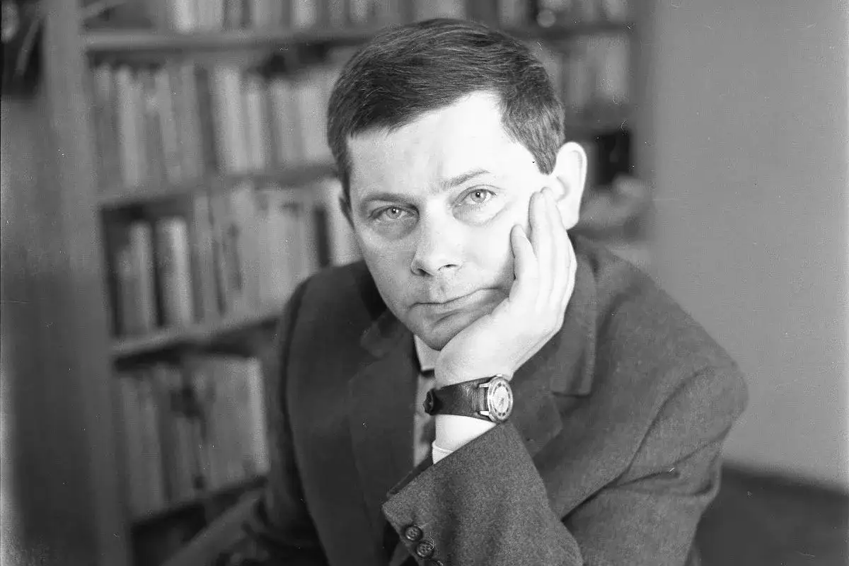Zbigniew Herbert, zamyślony, oparty o dłoń, na tle biblioteczki. Jego biografia to fascynująca podróż przez historię.